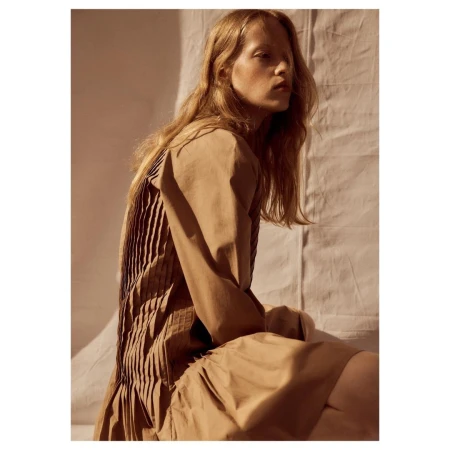 Model in a tan pleated dress seated against a wrinkled fabric background with warm sunlight