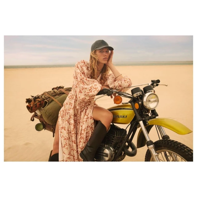 Woman wearing floral maxi dress and cap sits astride yellow vintage Kawasaki motorbike in desert landscape with horizon in the background.