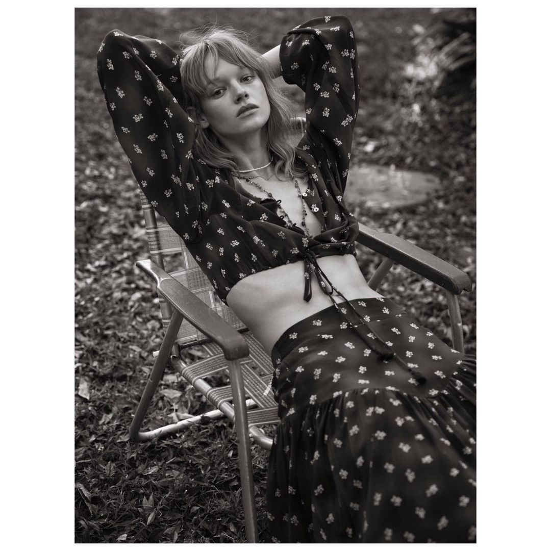 Black and white outdoor shot of a fair-skinned young woman in a floral crop top and skirt, sitting in a wooden chair, hands behind head.