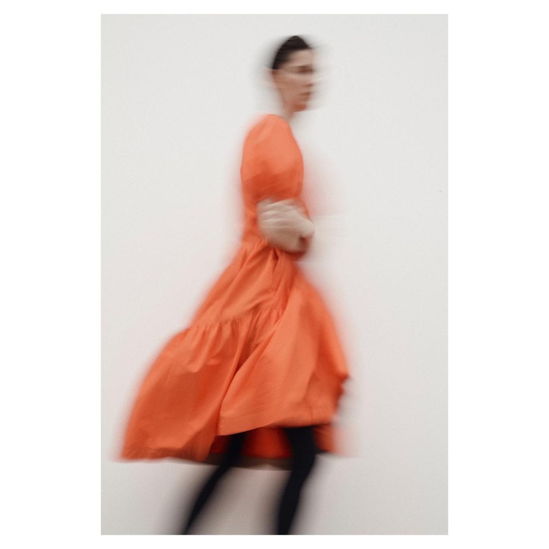 Blurred motion fashion shot of a young adult female wearing a tiered orange dress against a white wall in a studio setting.