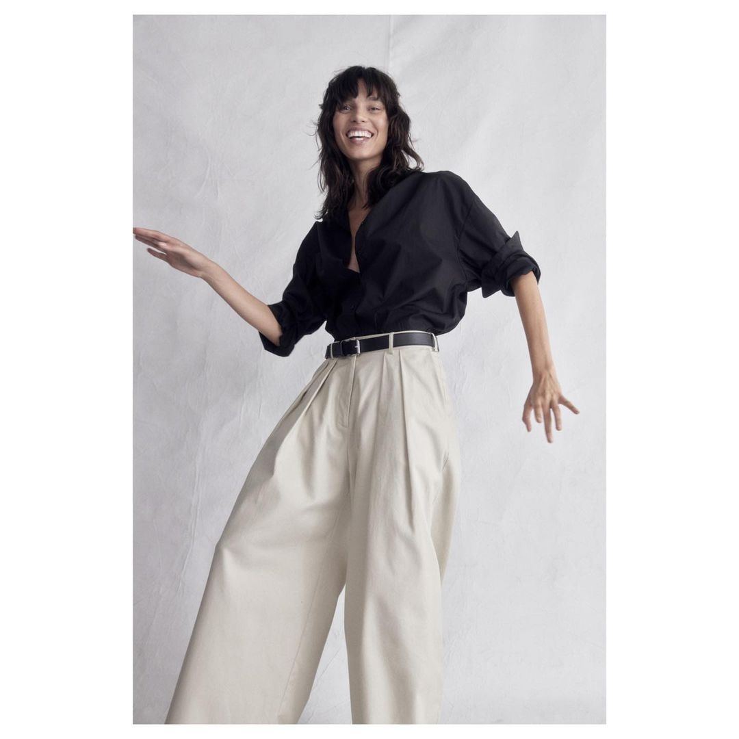 A smiling woman in a black button-down shirt and beige palazzo pants poses in a studio, exuding fashion-forward style and confidence.