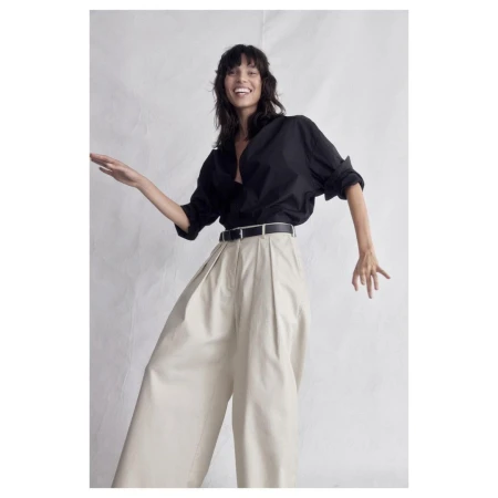 A smiling woman in a black button-down shirt and beige palazzo pants poses in a studio, exuding fashion-forward style and confidence.