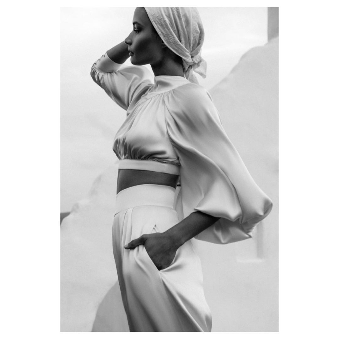 Monochrome studio shot of a woman in satin crop top and pants, wearing a headscarf, standing against a white wall, facing left in profile.