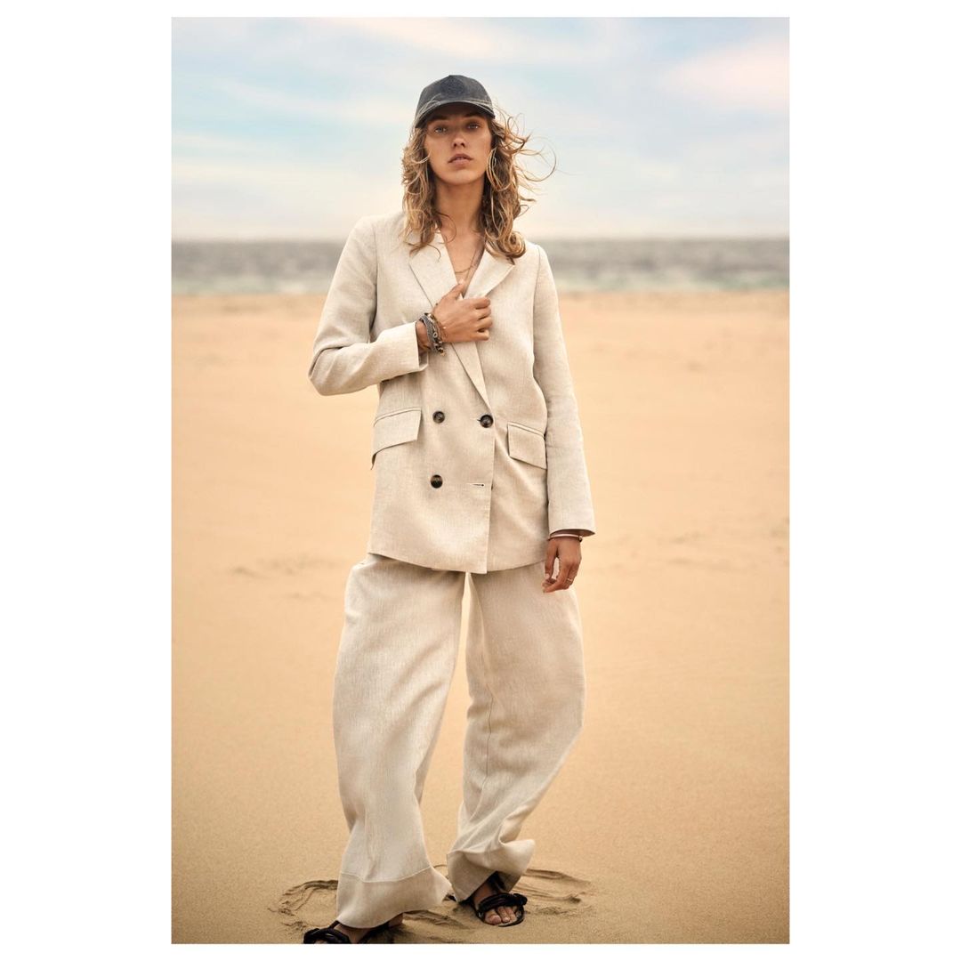 A woman in a linen suit and baseball cap poses on a sandy beach with the ocean in the background, showcasing fashion and editorial style.