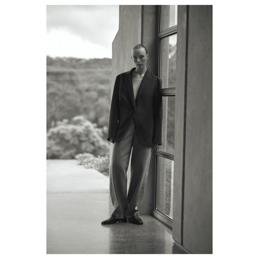 Black-and-white portrait of a woman in a blazer and wide-legged pants posing near a window with a nature background. Fashion editorial.