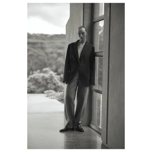 Black-and-white portrait of a woman in a blazer and wide-legged pants posing near a window with a nature background. Fashion editorial.