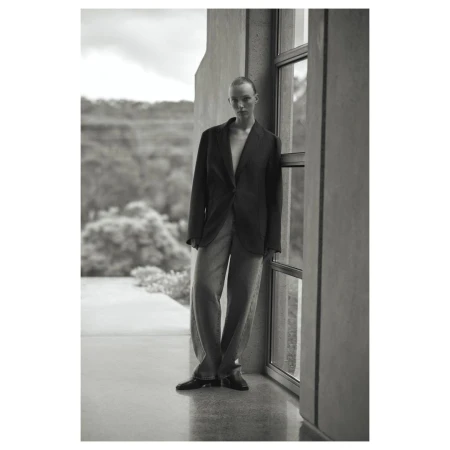Black-and-white portrait of a woman in a blazer and wide-legged pants posing near a window with a nature background. Fashion editorial.