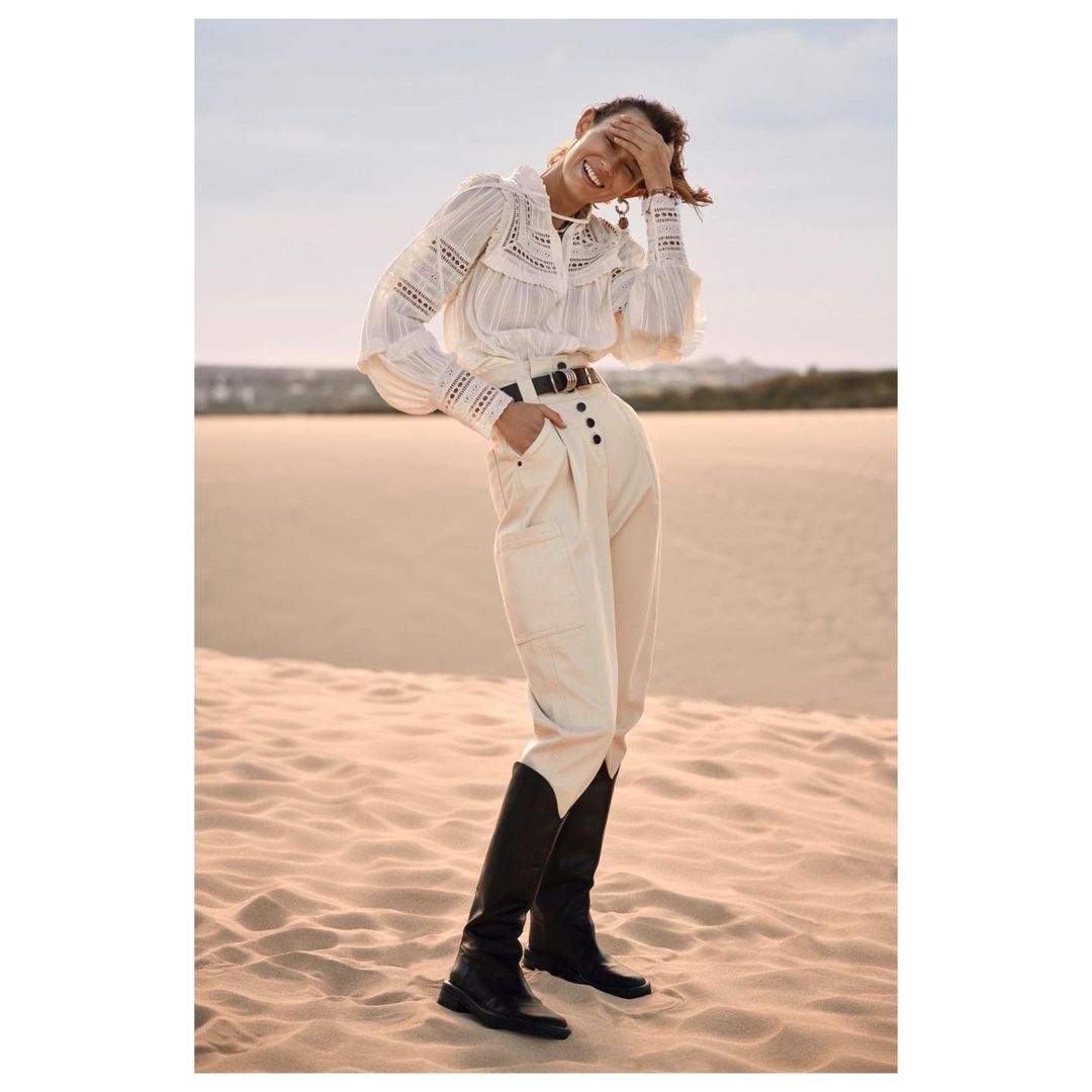 A woman in a white lace blouse and beige pants with black boots poses in a desert landscape in a fashion editorial style.