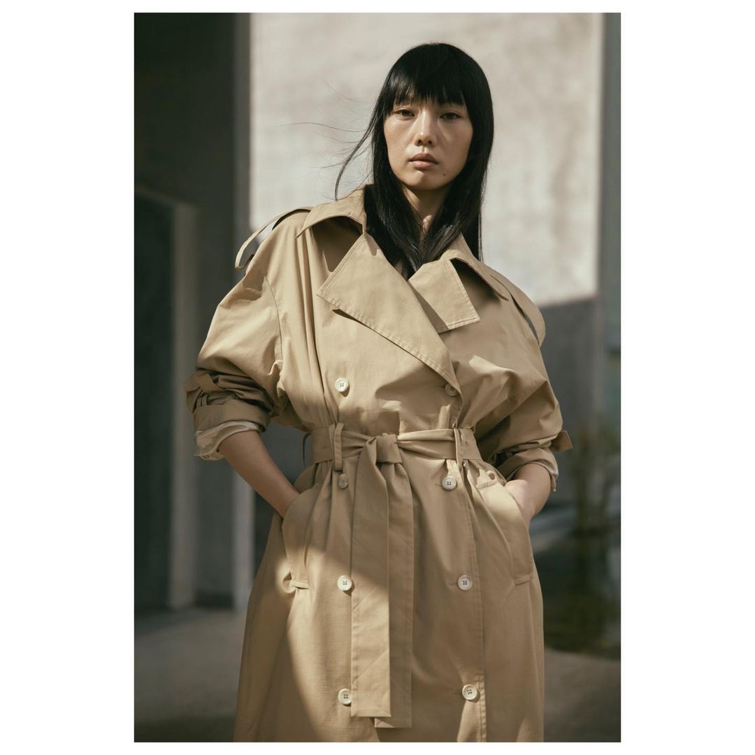 Portrait of an Asian woman wearing a tan trench coat with a belt, standing indoors with a neutral expression and dark hair.