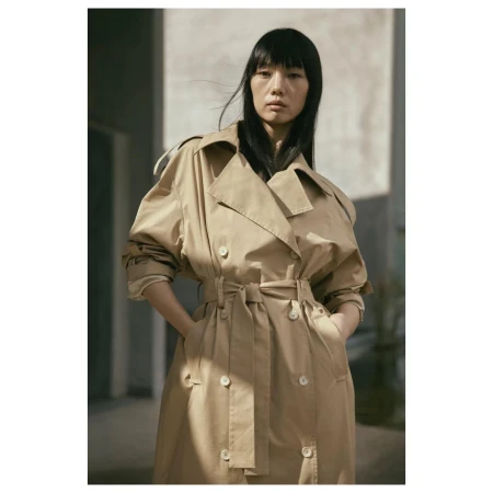 Portrait of an Asian woman wearing a tan trench coat with a belt, standing indoors with a neutral expression and dark hair.