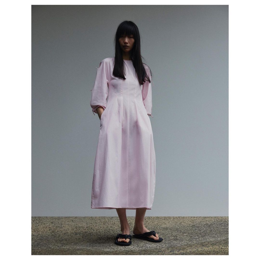 Full-length shot of a slender Asian woman in a pastel pink dress with black sandals, standing against a grey background.