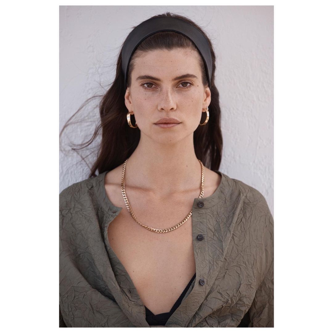 Portrait of a woman with dark hair and a black headband wearing a crinkled green blouse, gold hoop earrings and chain necklace against a white wall.