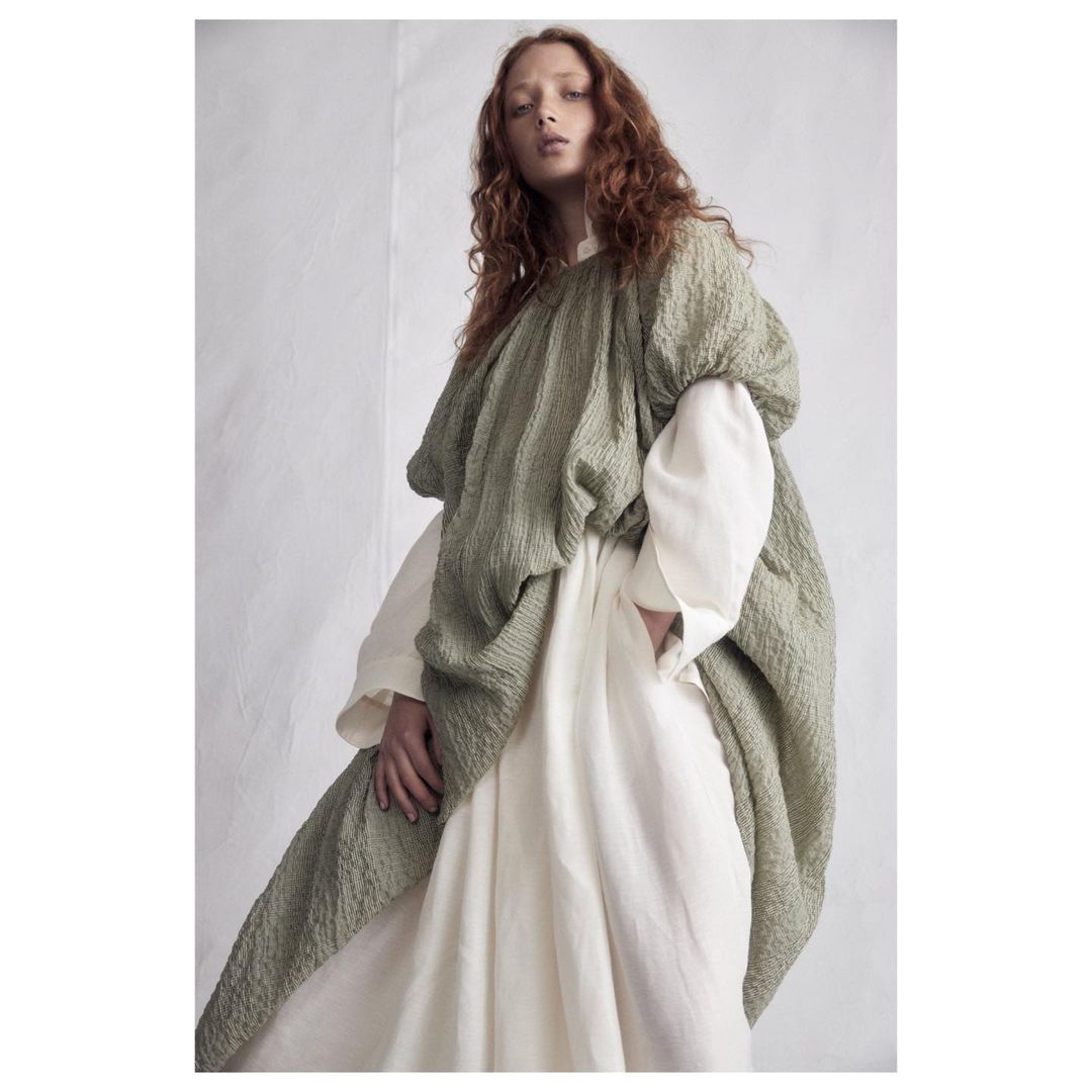 Model with long red hair wearing a textured olive green top over a white dress against a neutral background.