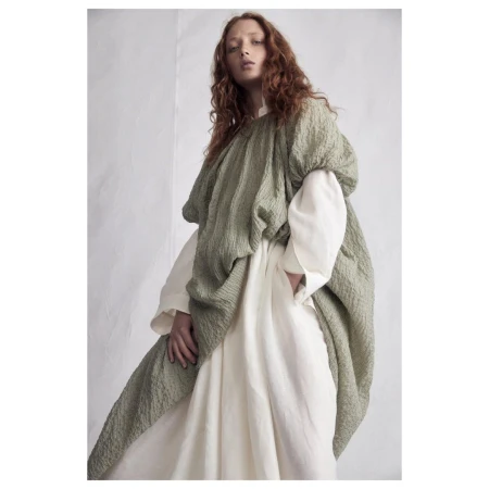 Model with long red hair wearing a textured olive green top over a white dress against a neutral background.