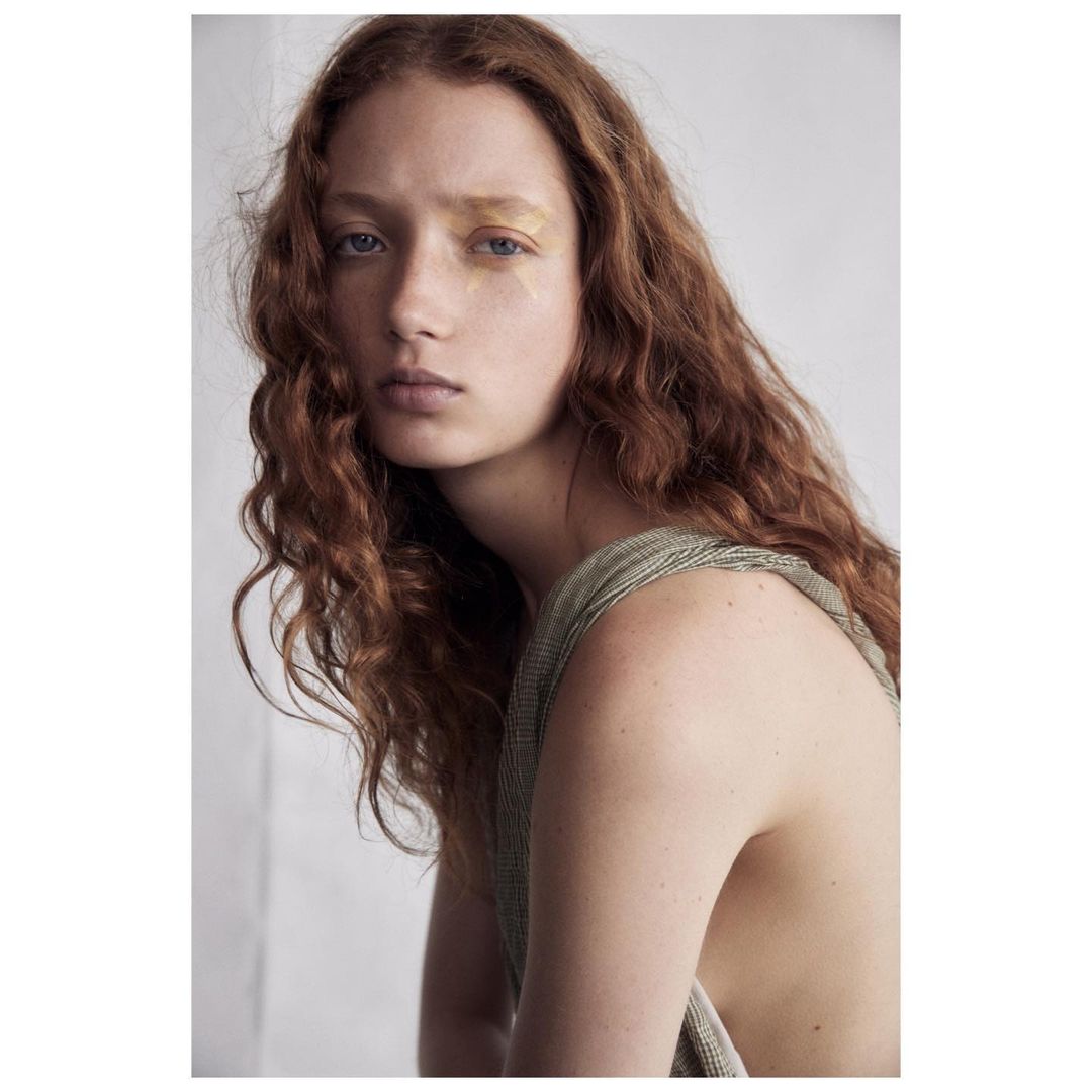 Portrait of a young woman with red hair and gold makeup, wearing a green top, against a neutral background.