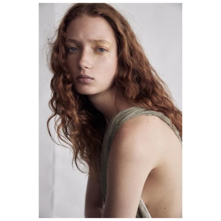 Portrait of a young woman with red hair and gold makeup, wearing a green top, against a neutral background.