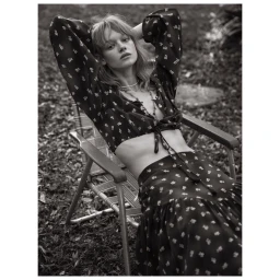 Monochrome fashion portrait of a female model seated in a folding lawn chair wearing a patterned long-sleeve crop top and skirt