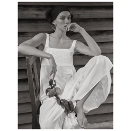 Model in white dress wearing black headscarf holding a peach branch while seated on a wooden chair.