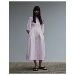 Model with long dark hair wearing a light pink midi dress and black sandals against a plain grey wall.