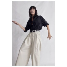 Smiling model wearing a black buttoned shirt and wide-legged beige trousers against a neutral textured studio background.