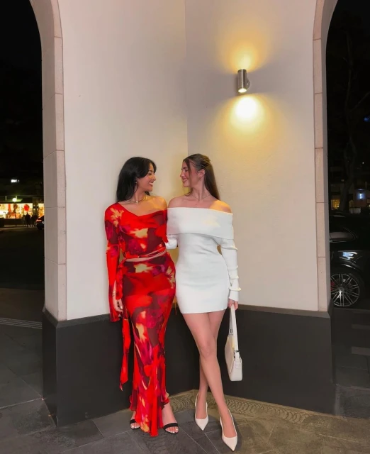 Two adult women pose outdoors at night, one in a red floral dress and the other in a short white dress. They are standing close together.