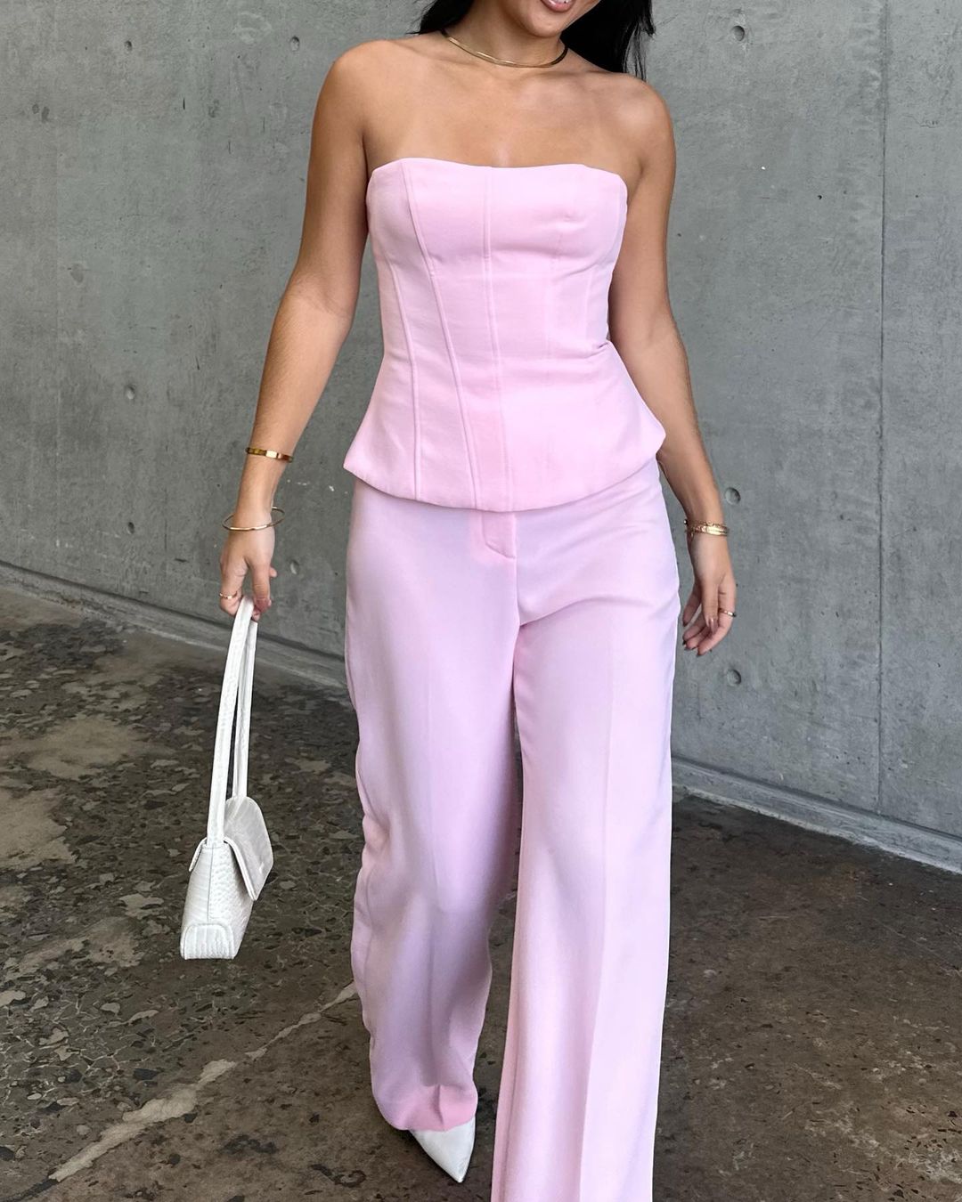 Woman in pink corset top and pants with white heels holding purse against concrete wall, fashion shoot, indoor setting, light colors.