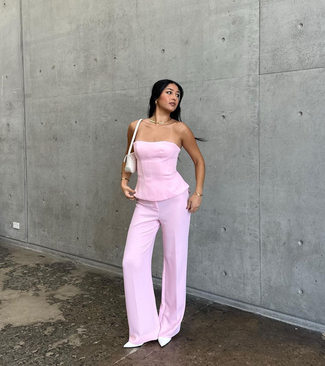 An Asian woman poses outdoors, wearing a light pink corset top and matching wide-leg pants, accessorized with a white purse and jewelry.