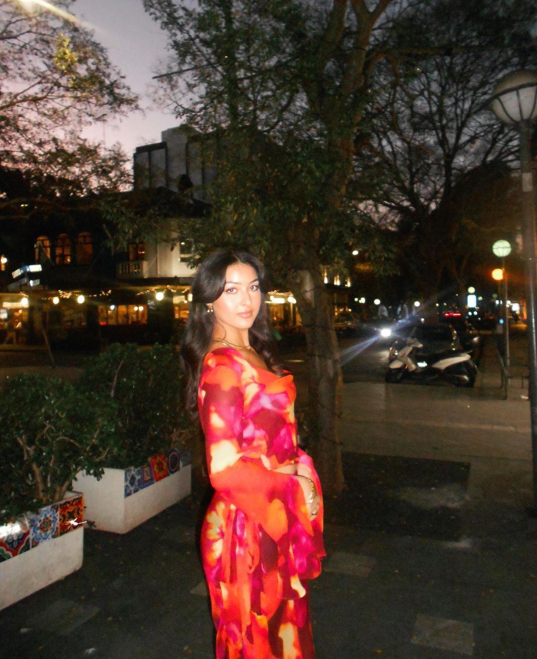 Attractive woman in a floral print maxi dress stands on a city street at dusk, with buildings and trees in the background.