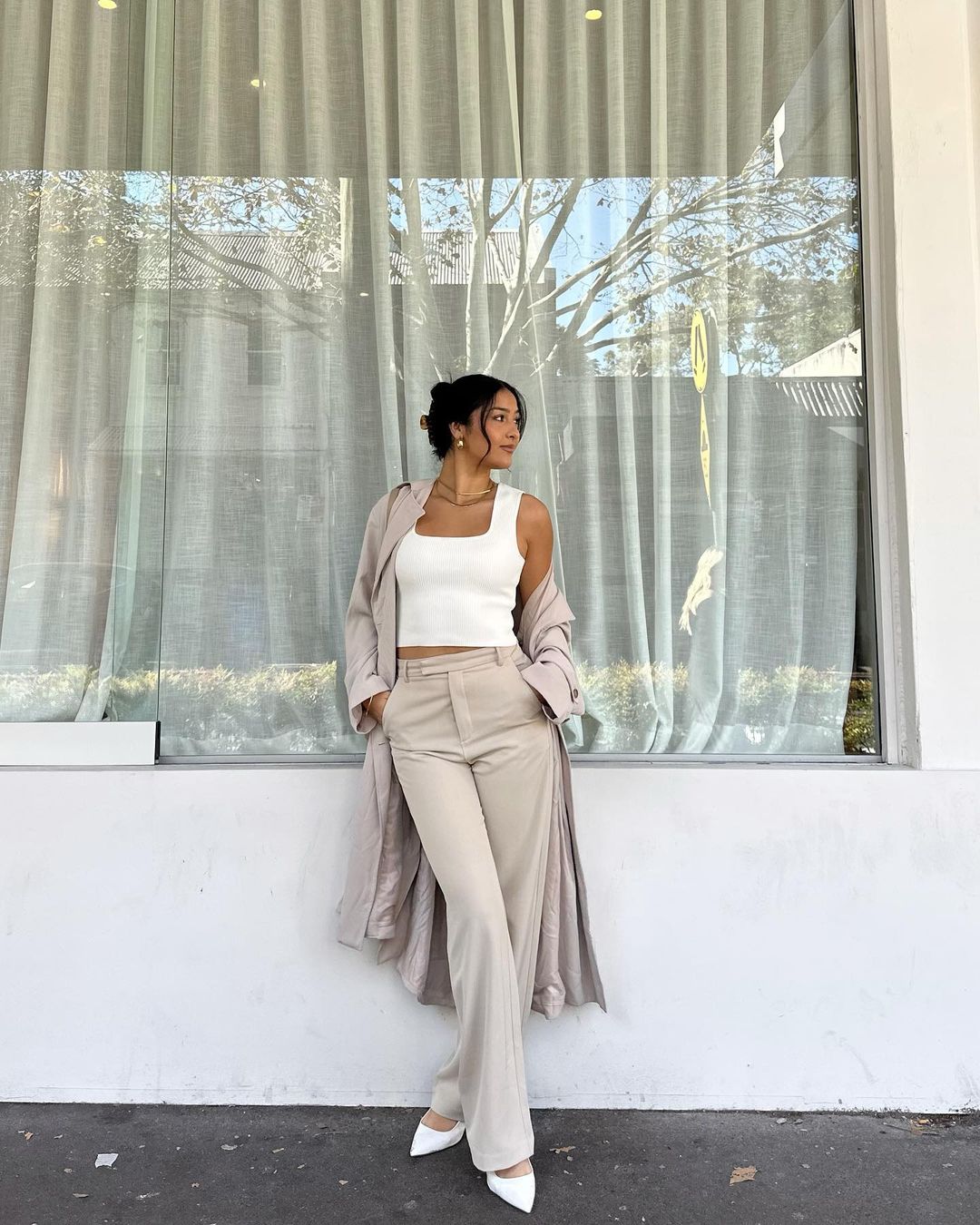 A stylish woman poses in front of a window, wearing a white top, beige pants, trench coat, and white heels in an indoor setting.