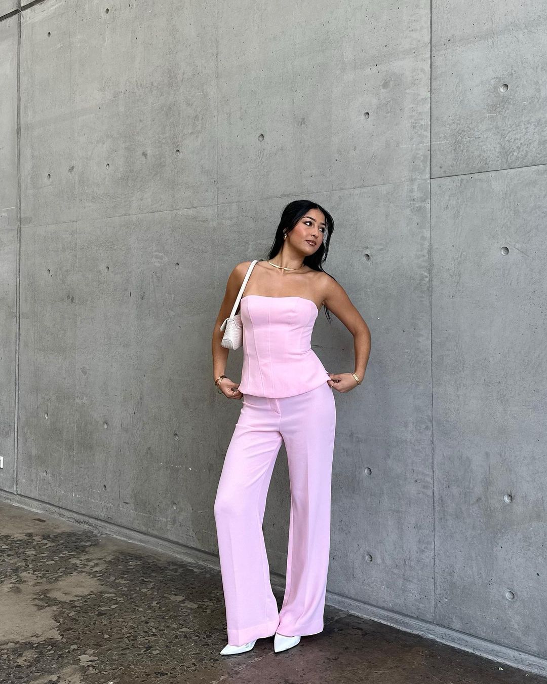 Woman in a pink pantsuit stands against a grey concrete wall, carrying a white purse. Fashion shoot with natural lighting.