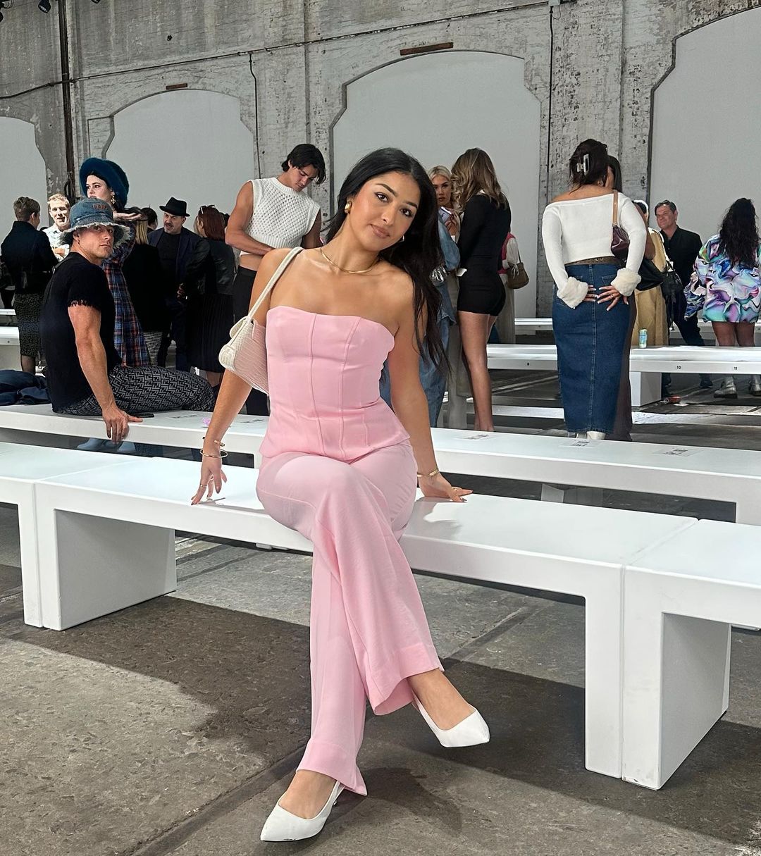 A stylish woman in a pink jumpsuit poses on a white bench at an indoor fashion event, surrounded by a crowd of onlookers and concrete floors.