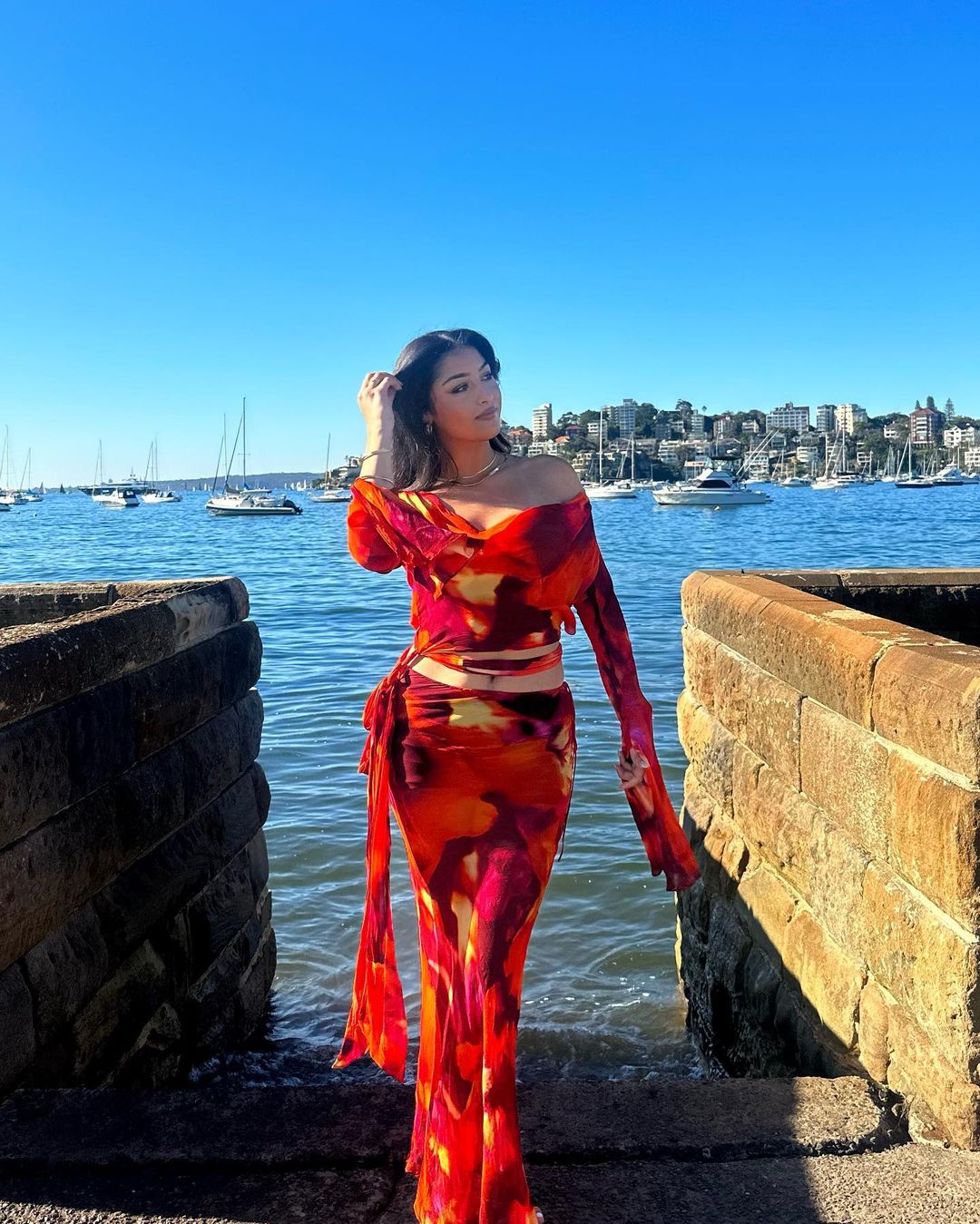 Young woman poses in vibrant tie-dye dress by ocean on sunny day. Yachts dot water with skyline and blue sky backdrop.