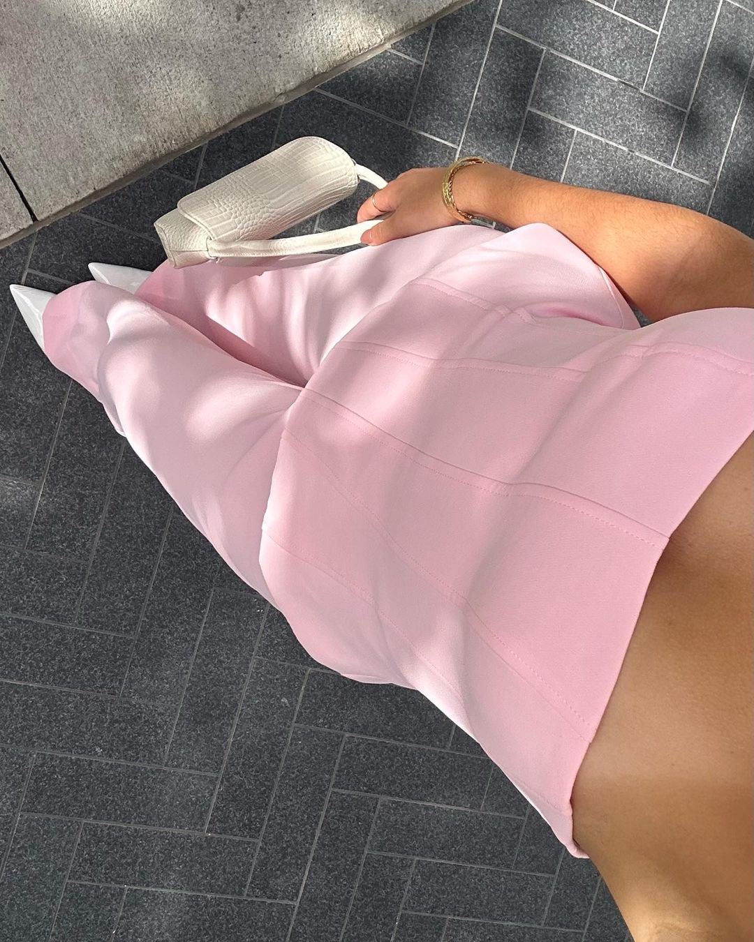 Woman wearing a pink dress and white heels, holding a white purse, stands on a patterned grey surface in natural sunlight.