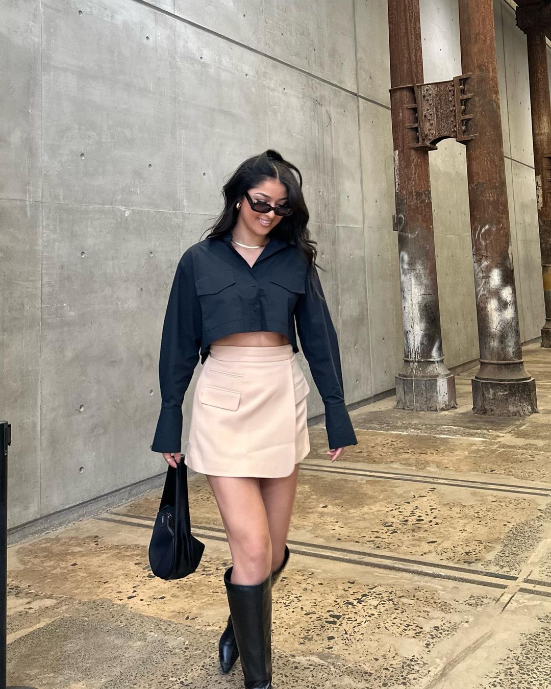 A stylish adult female poses confidently in a cropped black top, beige skirt, and black boots against a concrete and weathered column background.