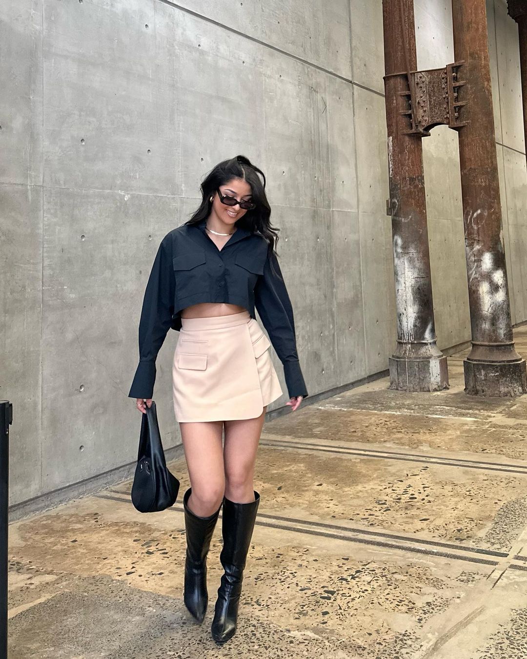 A woman in a black crop top, beige skirt, and black boots walks along a concrete walkway near pillars, wearing sunglasses and carrying a black bag.