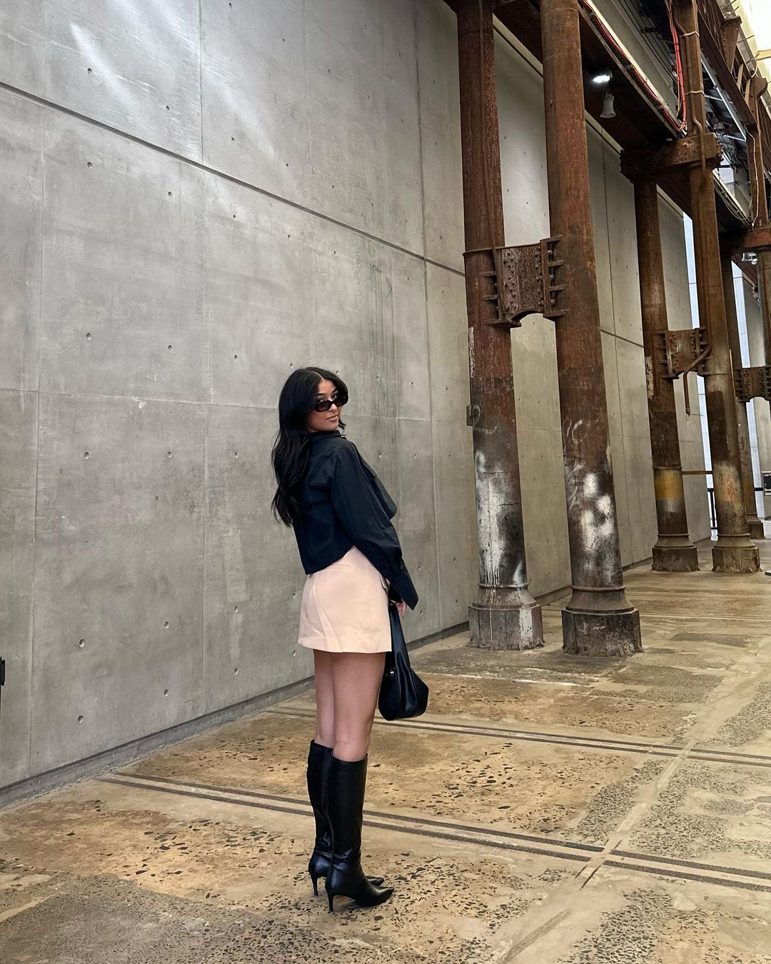 A woman in a black jacket, beige skirt, and black boots stands in an industrial space with concrete walls and weathered metal columns.