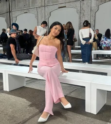 A woman in a pink strapless jumpsuit and white heels sits on a white bench at an event, captured by Sarah Bada.