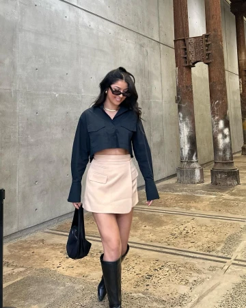 Woman in black crop shirt, beige mini skirt, and black boots walking near industrial concrete columns