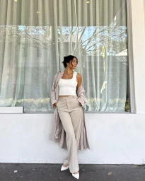 Woman wearing a white top and beige trousers leaning against a wall beneath large windows with sheer curtains.