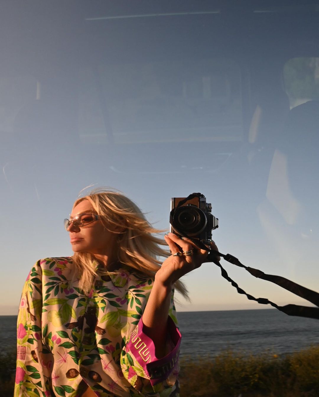 Adult female taking a selfie with an analog camera in front of a waterscape during daytime, wearing a patterned top and sunglasses.