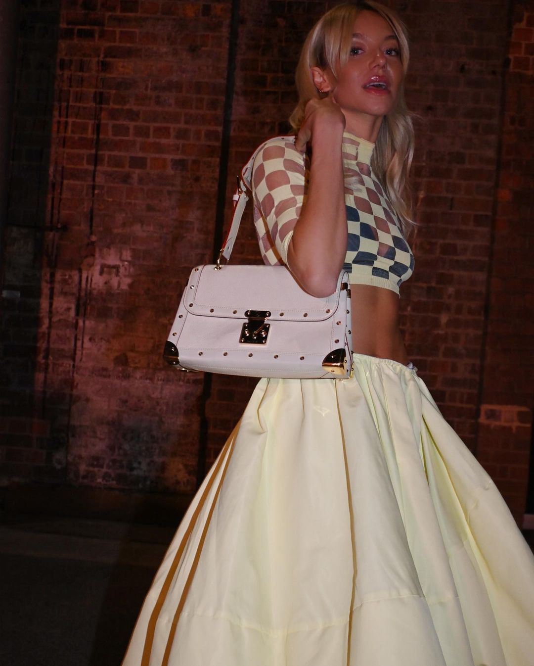 Woman in checkered top and yellow skirt carries a white purse against a brick background in a fashion setting. Blonde hair.