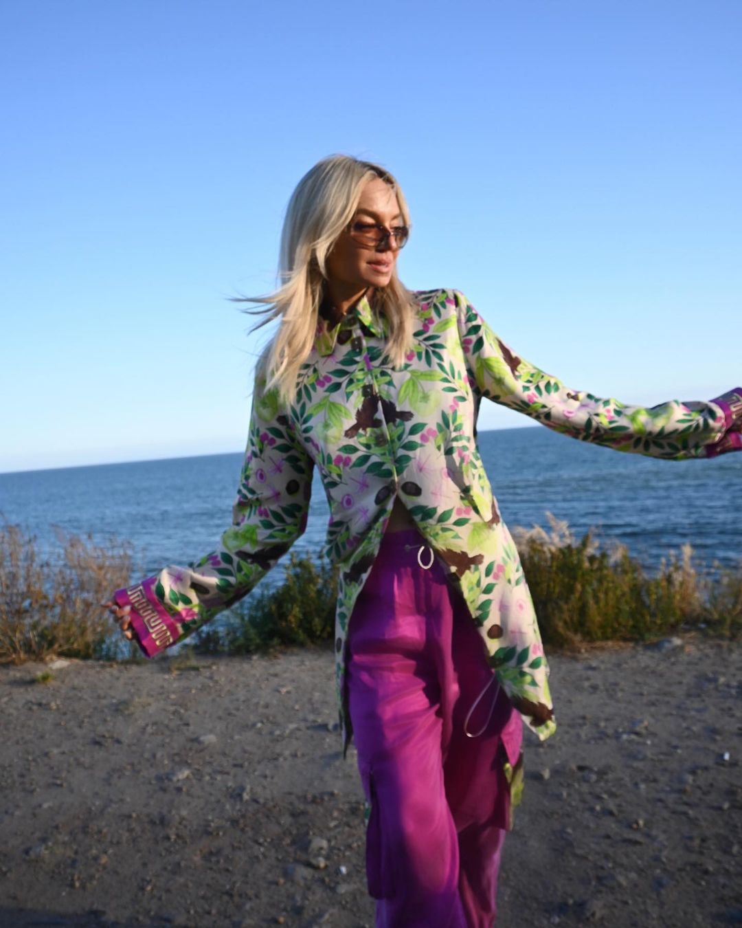 Woman with blonde hair and sunglasses standing on a beach, wearing a floral shirt and pink pants with the ocean and sky in the background.