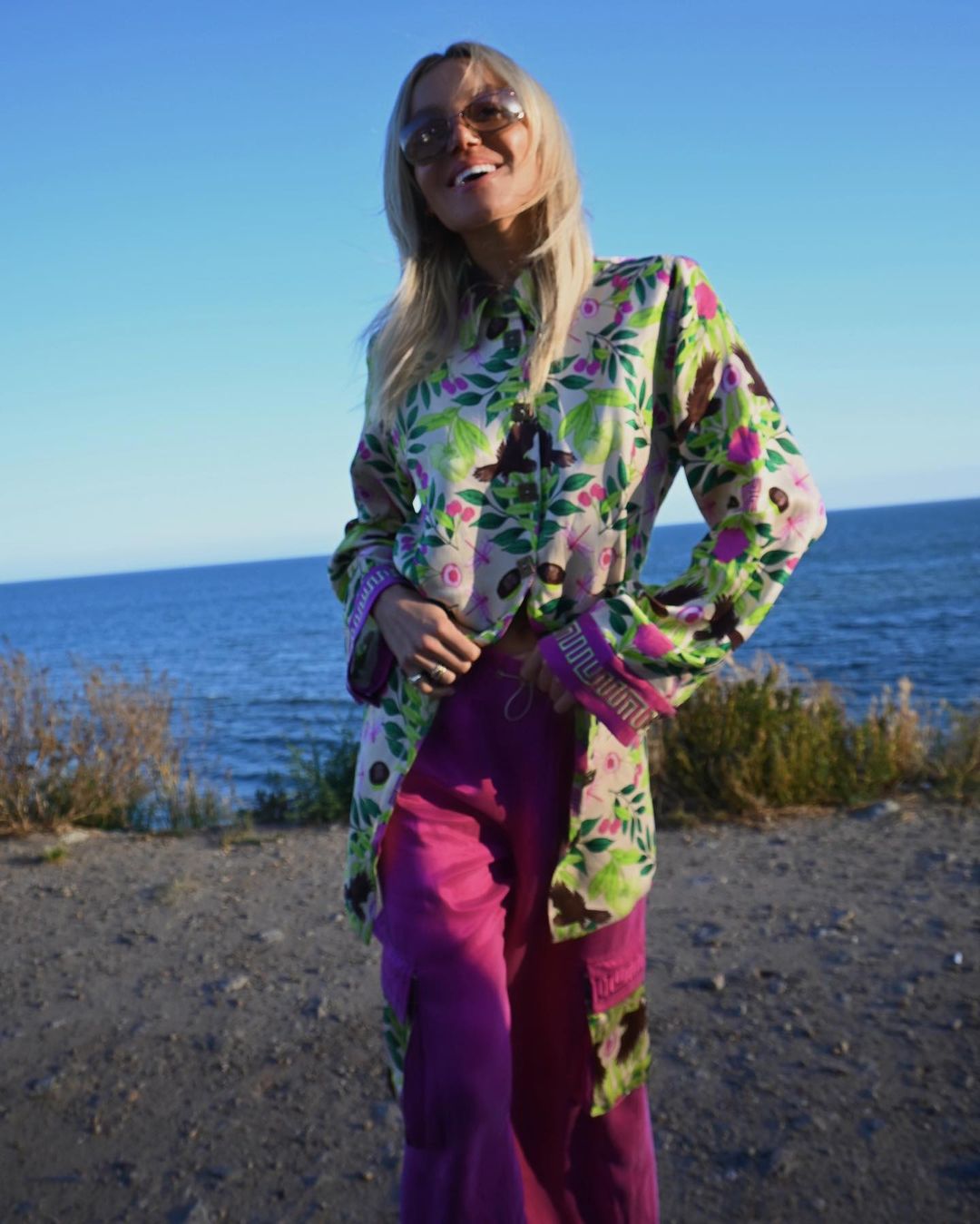 A smiling woman with blonde hair in a floral shirt and purple pants stands outside near the ocean under a clear, bright blue sky.