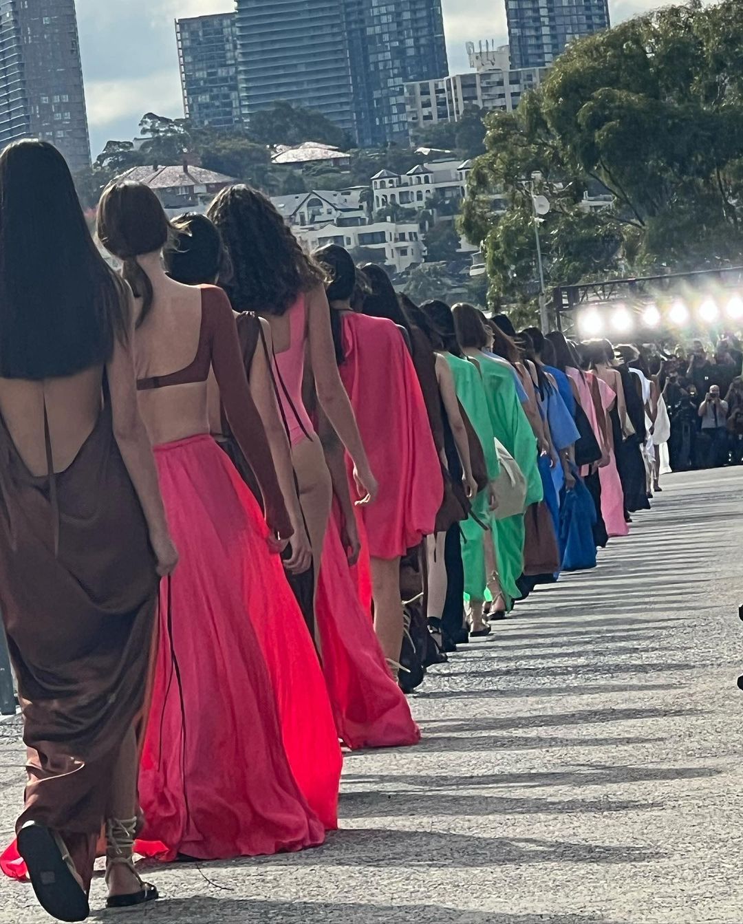 Fashion show featuring models walking down an outdoor runway wearing colorful dresses with an urban skyline and crowd in the background.