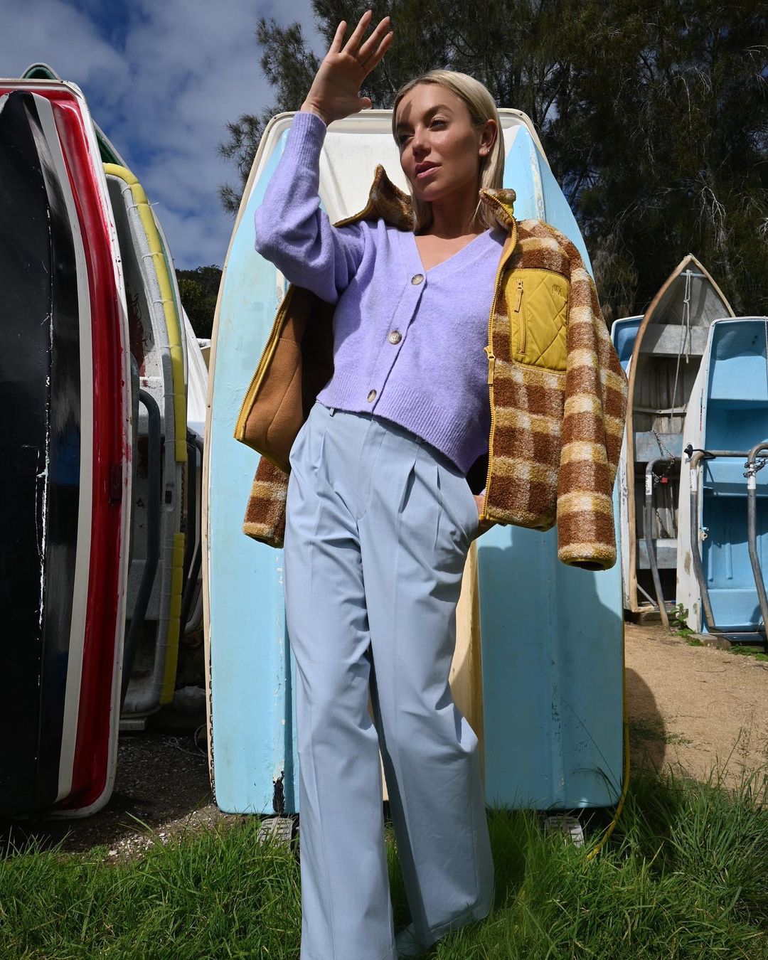 A woman in a lavender sweater and plaid jacket stands among colorful rowboats on green grass, shielding her eyes from sunlight.