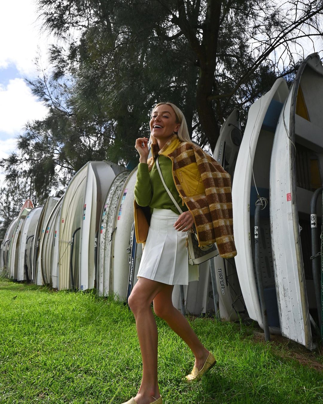 A woman strolls in a green plaid jacket and white skirt past stacked boats on green grass on a sunny day, smiling and happy.