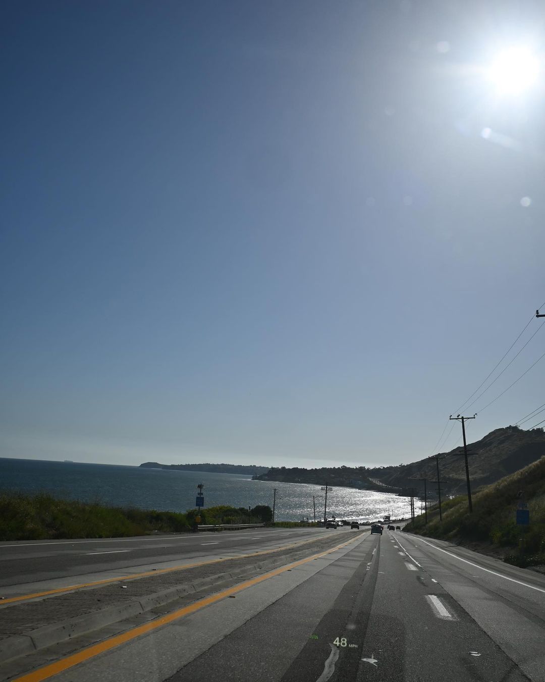 Coastal road with asphalt, yellow lane markings, ocean, and hills under a bright blue sky and intense sun, showcasing a scenic drive.