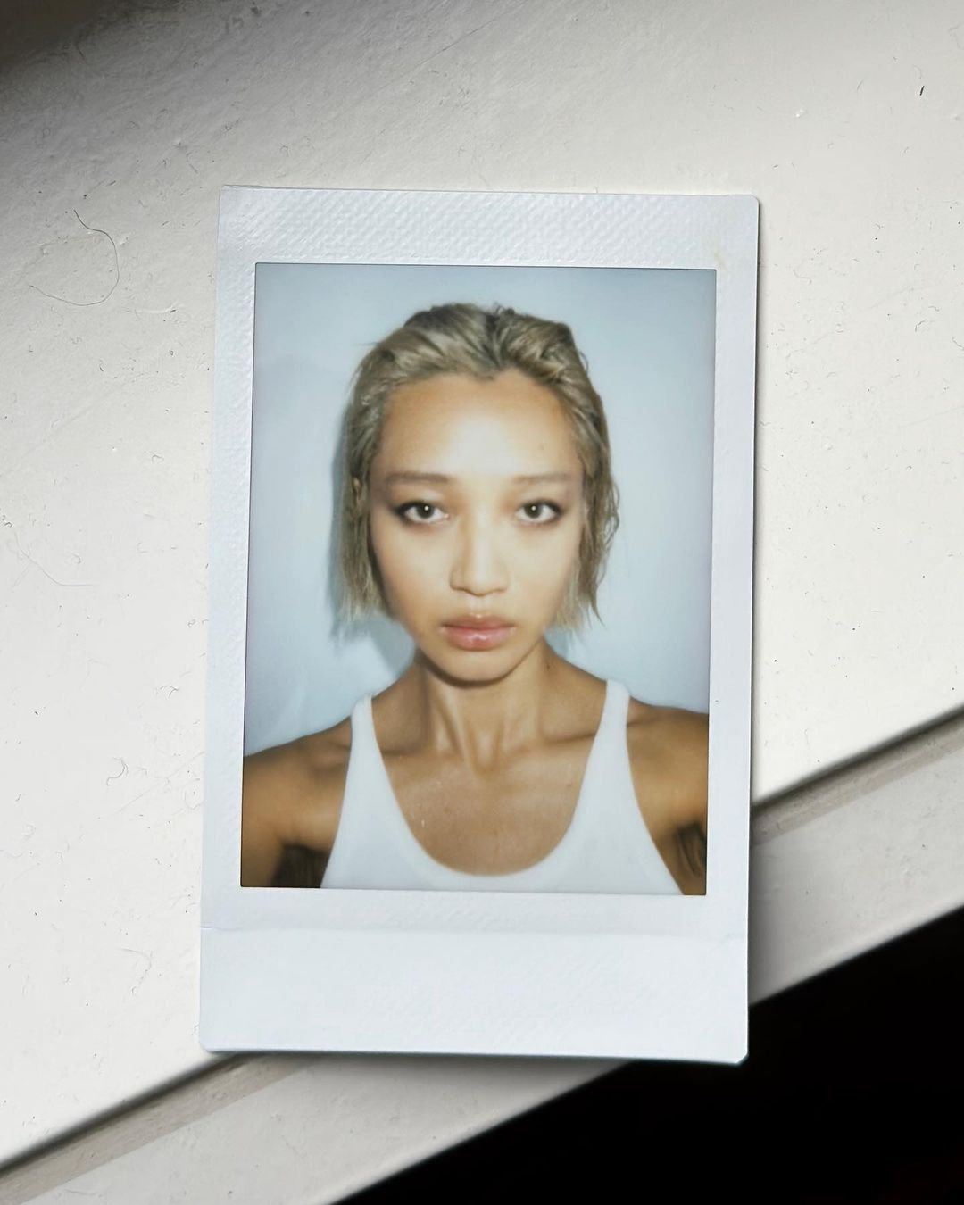 Portrait of a young woman with light brown hair, wearing a white tank top, with fair skin tone, in a polaroid-style photograph.