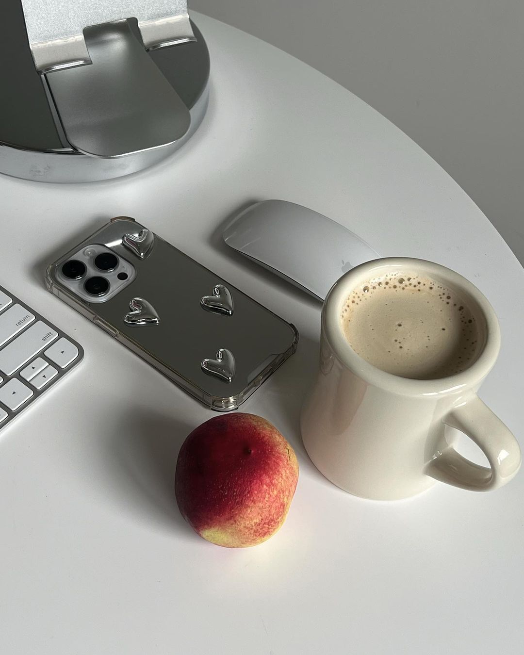 Overhead shot of a coffee mug, smartphone, peach, and keyboard on a white table, creating a minimalist and clean aesthetic.