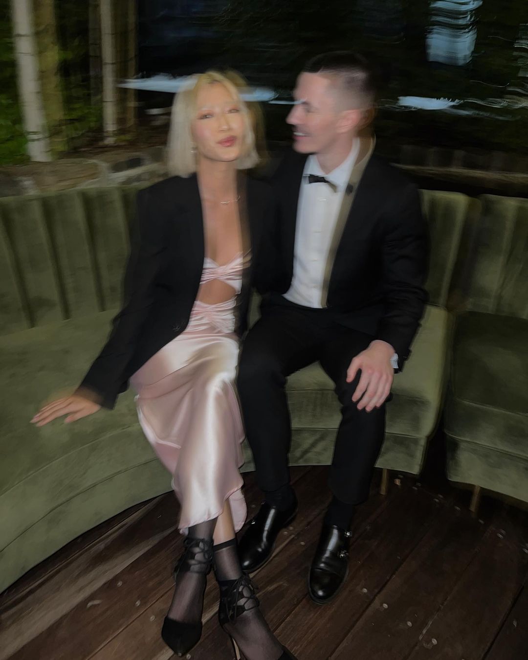 Well-dressed couple poses indoors on a green velvet couch. The woman wears a pink dress and the man wears a black suit with a bow tie.